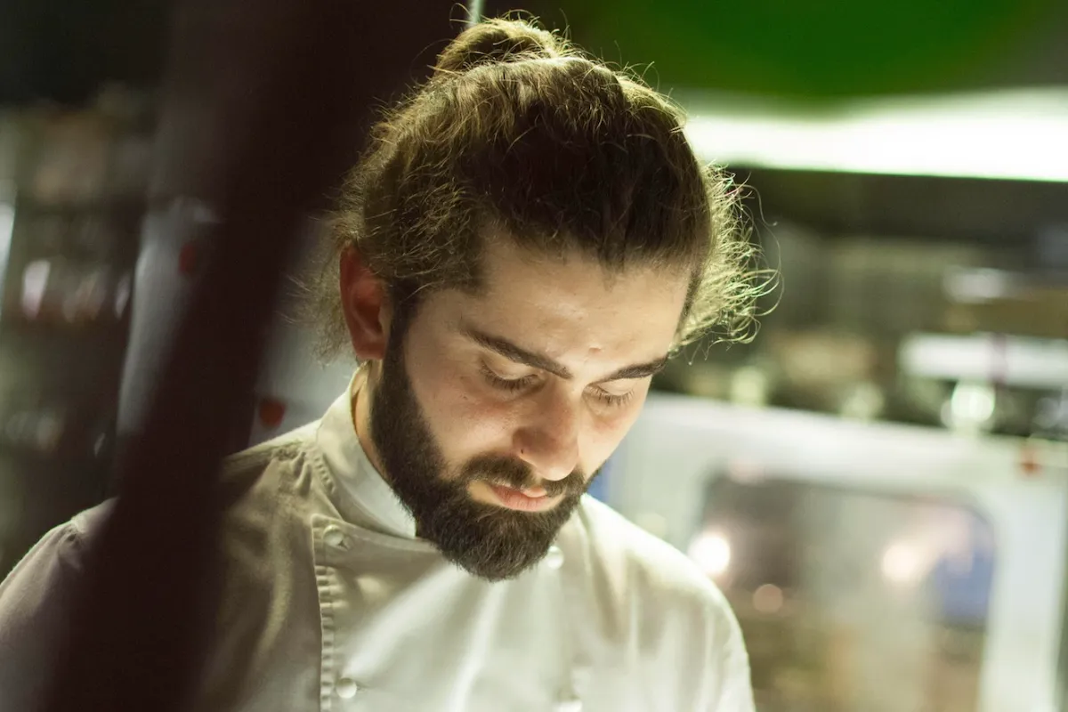 Beyaz şef ceketini giymiş, saçları topuz yapılmış ve gür sakallı Şef İbrahim Tunç, sürdürülebilir gastronomi anlayışıyla oluşturduğu eşsiz lezzet rotalarını ve mutfak felsefesini düşünürcesine gözleri kapalı veya aşağı doğru odaklanmış bir şekilde görünüyor.