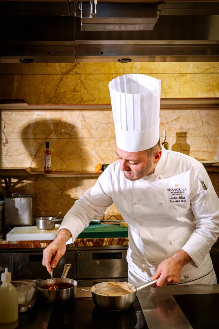 Shangri-La Bosphorus İstanbul'un Executive Şefi Şevket Cihan, beyaz şef ceketi ve klasik şapkasıyla modern bir mutfakta, mermer dokulu arka planın ve paslanmaz çelik davlumbazın önünde, dikkatle iki tencerede yemek karıştırarak hünerini sergiliyor.