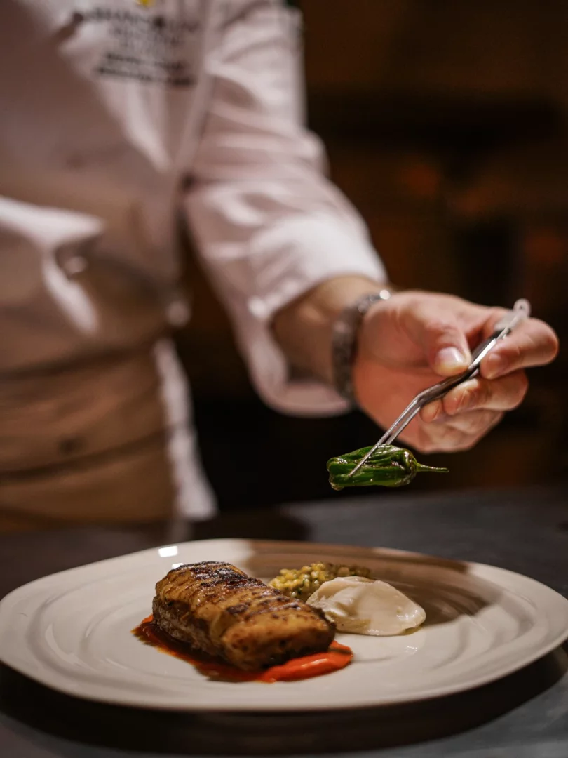 Fine dining deneyiminin inceliğini yansıtan bu görselde, bir şefin beyaz bir tabağa özenle yerleştirilmiş ızgara ana yemeğin, renkli püreler ve taneli garnitürlerle bütünleşen zarif tabak sunumuna maşa yardımıyla küçük bir yeşil biber eklediği an vurgulanıyor.