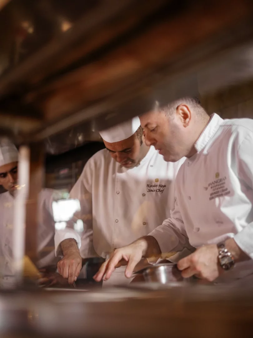 Şef Şevket Cihan, Shangri-La Bosphorus İstanbul mutfağında beyaz şef ceketleri giymiş ekibiyle paslanmaz çelik bir tezgah üzerinde özenle çalışırken, farm-to-table yaklaşımının ve yerel ürünlerin değerini yansıtan bir mutfak atmosferinde detaylara odaklanıyor.