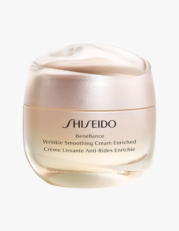 Beyaz bir arka plan üzerinde, zarif tasarımlı, şeffaf kapaklı ve hafif şeftali tonlarına sahip Shiseido Benefiance Wrinkle Smoothing Cream Enriched yaşlanma karşıtı krem kavanozu, ürünün lüks ve etkili yapısını vurguluyor.