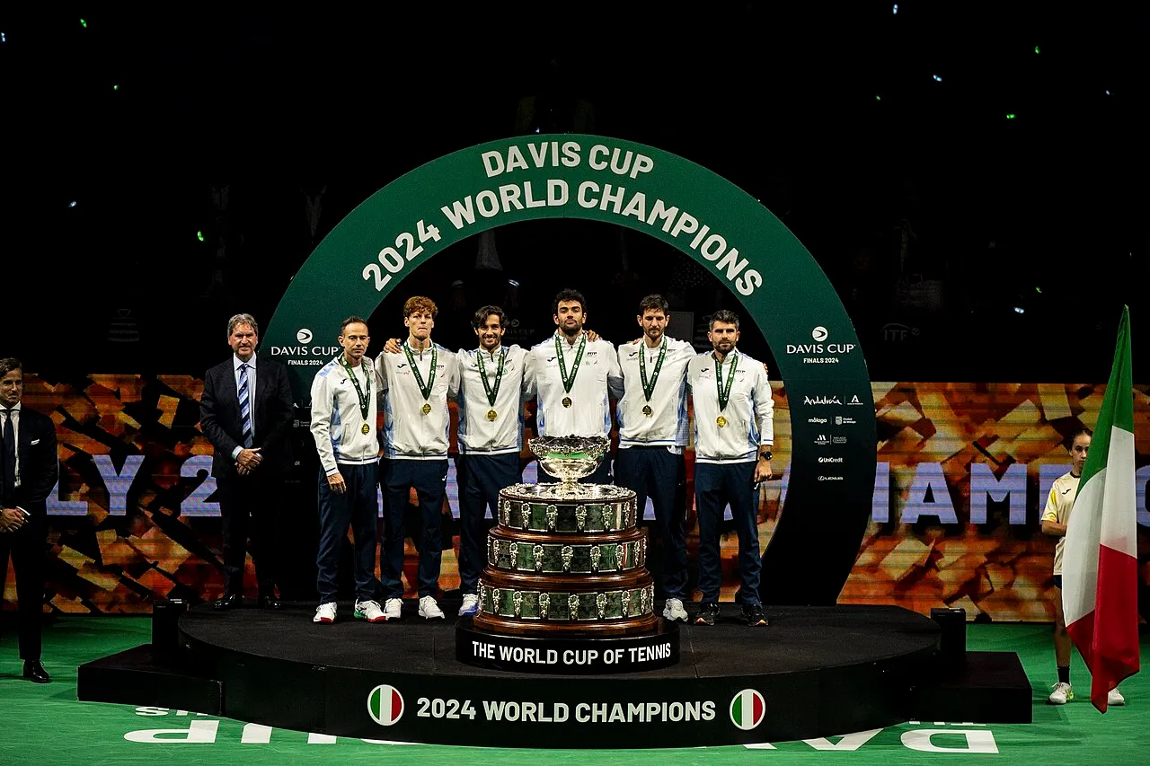 Davis Kupası 2024 Dünya Şampiyonası'nı kazanan İtalya tenis takımı üyeleri, podyumda parıldayan gümüş kupanın arkasında, boyunlarında altın madalyalarla gururla sıralanmış, zaferlerini kutluyor; arka plandaki yeşil kemerde DAVIS CUP 2024 WORLD CHAMPIONS yazısı ve sağdaki İtalyan bayrağı bu tarihi anı pekiştiriyor.
