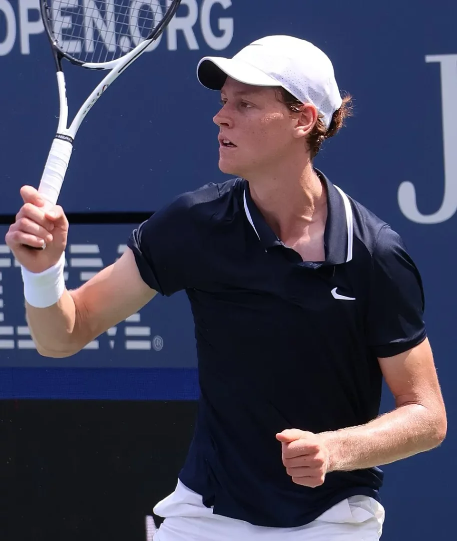 Jannik Sinner, 2024 US Open turnuvasında, mavi bir kort zemininde, beyaz şapkası, Nike logolu lacivert tişörtü ve beyaz bilekliğiyle, tenis raketini iki eliyle kavrayarak topa vurmaya hazırlanırken veya vuruş sonrası anın odağında, ciddi ve konsantre bir yüz ifadesiyle görülüyor.