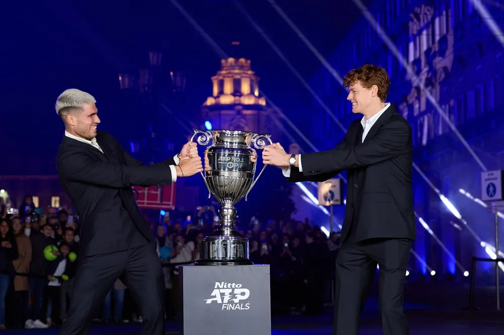 Carlos Alcaraz ve Jannik Sinner, Nitto ATP Finals podyumunda parlak gümüş bir kupayı aynı anda tutarak neşeyle çekişirken, mavi ışıklarla aydınlatılmış arka planda kalabalık seçiliyor ve bu an onların tenis rekabetini eğlenceli bir biçimde yansıtıyor.