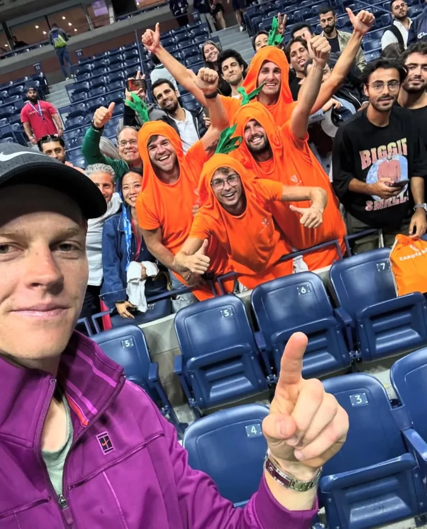 Jannik Sinner'ın tribün koltuklarında mor ceketiyle selfie çekerken işaret parmağını havaya kaldırdığı, arkasında turuncu havuç kostümleri giymiş Carota Boys hayran grubunun coşkuyla gülümsediği ve başparmaklarını yukarıda tuttuğu canlı bir an.