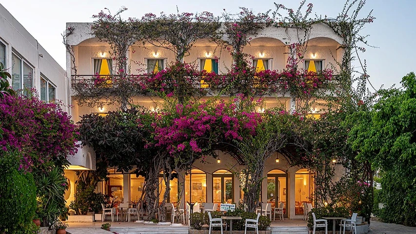 Skala Hotel Patmos'un mor begonviller ve yemyeşil sarmaşıklarla bezeli, beyaz badanalı üç katlı binası, akşam ışıklarıyla parlayan pencereleri ve balkonlarıyla sıcak bir Akdeniz atmosferi sunarken, zemin katta yer alan açık hava oturma alanında şık beyaz masalar ve sandalyeler dikkat çekiyor.