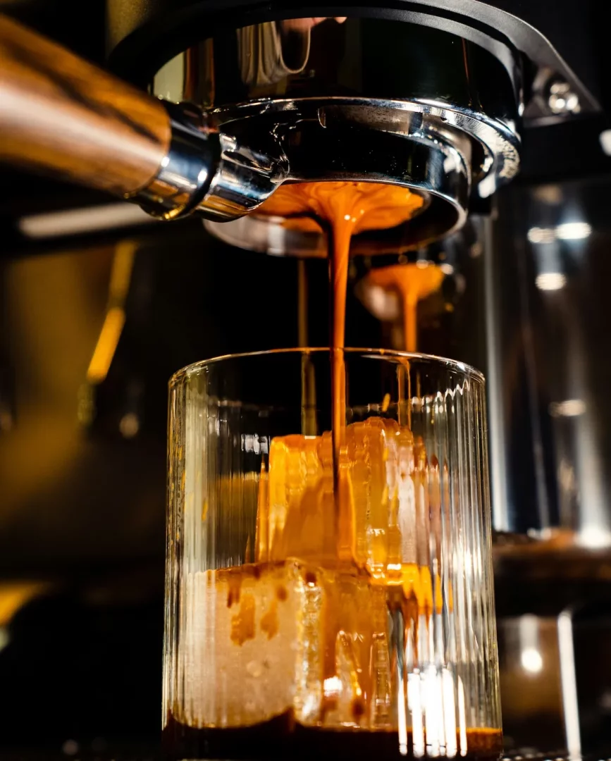 Yoğun kıvamlı espresso, bir espresso makinesinden buz küpleriyle dolu çizgili şeffaf bir bardağa dökülürken, soğuk kahve hazırlığının önemli bir aşaması olan buzların üzerine akışını ve bardağın dibinde koyu bir katman oluşturmaya başladığını gösteriyor.
