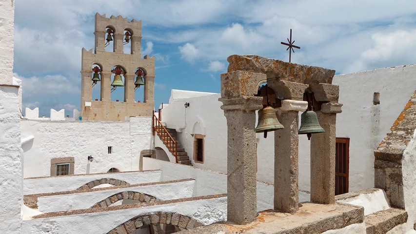 Patmos'taki Aziz Yuhanna Manastırı'nda, beyaz badanalı duvarlar arasında yükselen, gökyüzüne uzanan taş çan kulesinin ve ön plandaki iki çanlı küçük taş yapının, mavi ve bulutlu bir gökyüzü altında gösterişli bir görüntüsü.