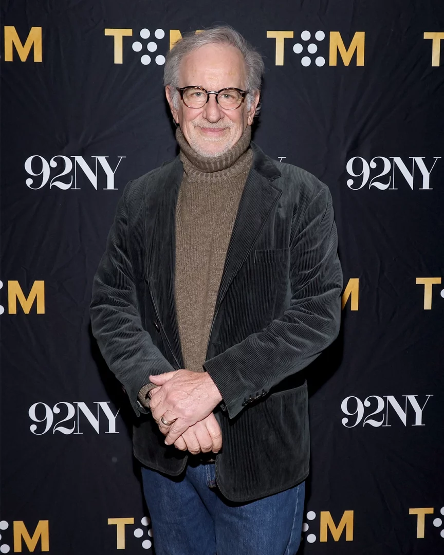 Gri saçlı, gözlüklü ve gülümseyen ünlü yönetmen Steven Spielberg, kahverengi boğazlı kazağı ve koyu renk kadife ceketiyle elleri önde kenetlenmiş şekilde, 92NY ve T:M logolu siyah bir panelin önünde duruyor.