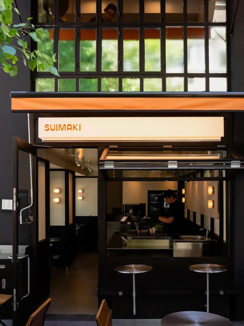 Suimaki isimli, koyu renkli modern bir işletmenin dış cephesinde, turuncu bir saçak altında beyaz panelde marka adı yazarken, açık servis bölümünde bir çalışan tezgahın arkasında görülüyor; üst kat pencerelerinden bakan bir kişi ve dışarıda yuvarlak metal masalar, çağdaş bir restoran veya kafenin hareketli ambiyansını sergiliyor.