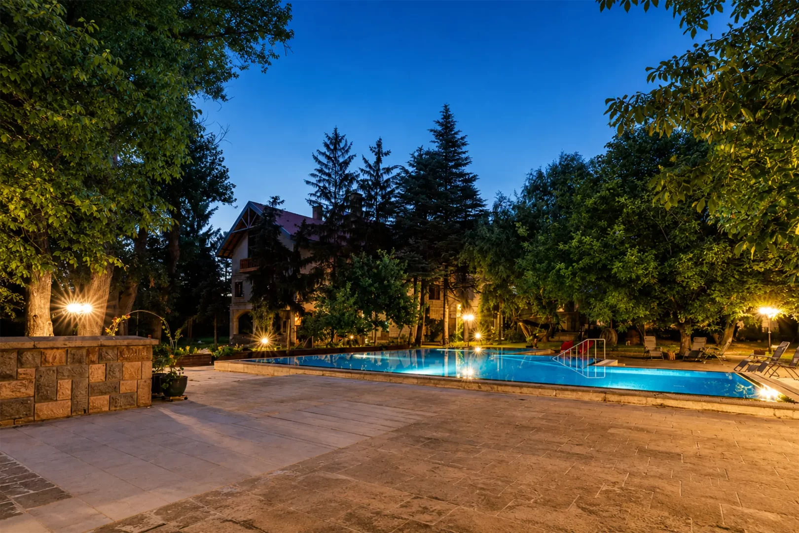 Alacakaranlıkta çekilmiş bu geniş açılı fotoğrafta, çevresi ağaçlar ve ışıklarla aydınlatılmış taş döşeli geniş bir terasın ortasında yer alan pırıl pırıl mavi bir açık yüzme havuzu ve arka planda kiremit çatılı geleneksel bir bağ evi huzurlu bir atmosfer sunuyor.