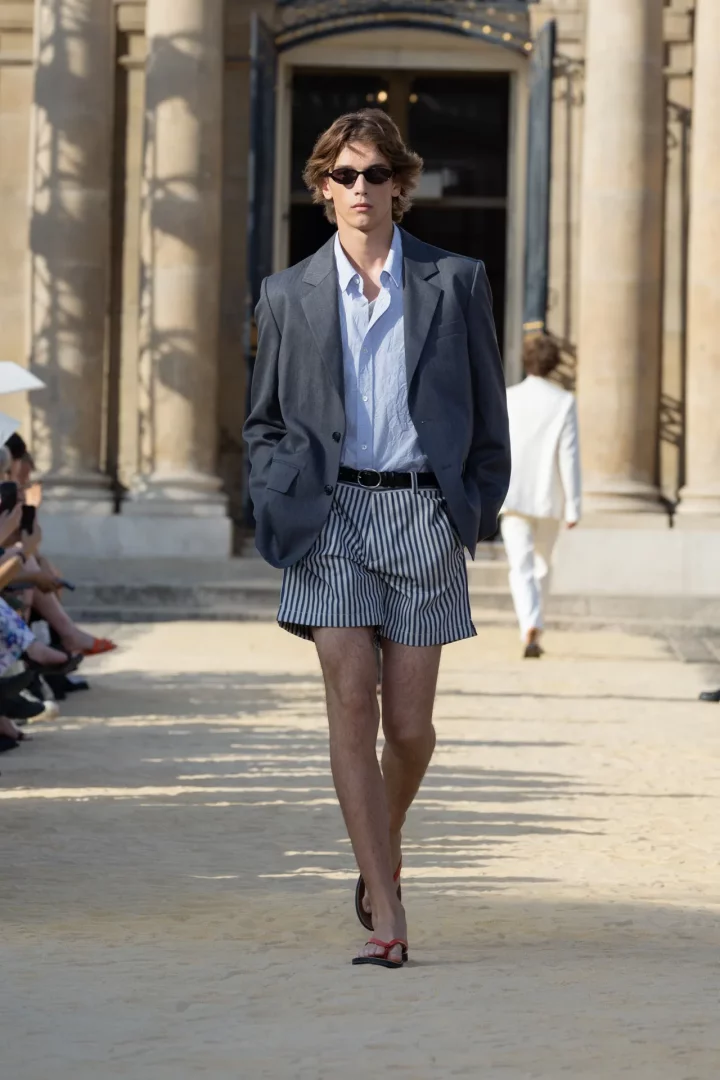2027 İlkbahar Paris Moda Haftası defilesinde, dalgalı kahverengi saçlı bir erkek model, gri blazer ceket, açık mavi gömlek, çizgili şort ve kırmızı parmak arası terlikleriyle güneş gözlüğü takarak klasik bir binanın önündeki açık hava podyumunda kendinden emin bir şekilde yürüyor.