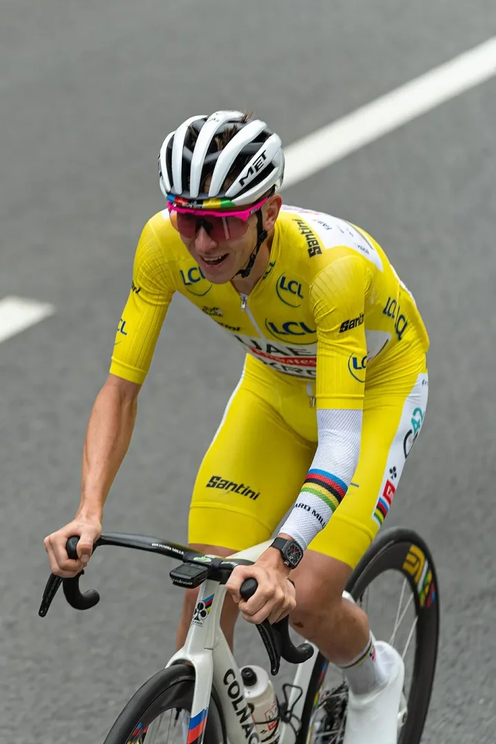 Tour de France'ın liderlik sarı mayosuyla beyaz Colnago bisikleti üzerinde pedal çeviren Tadej Pogačar, yüzündeki neşeli gülümsemesiyle, beyaz MET kaskı, pembe çerçeveli güneş gözlükleri ve kolundaki dünya şampiyonu gökkuşağı şeritleri ile yolda ilerliyor.