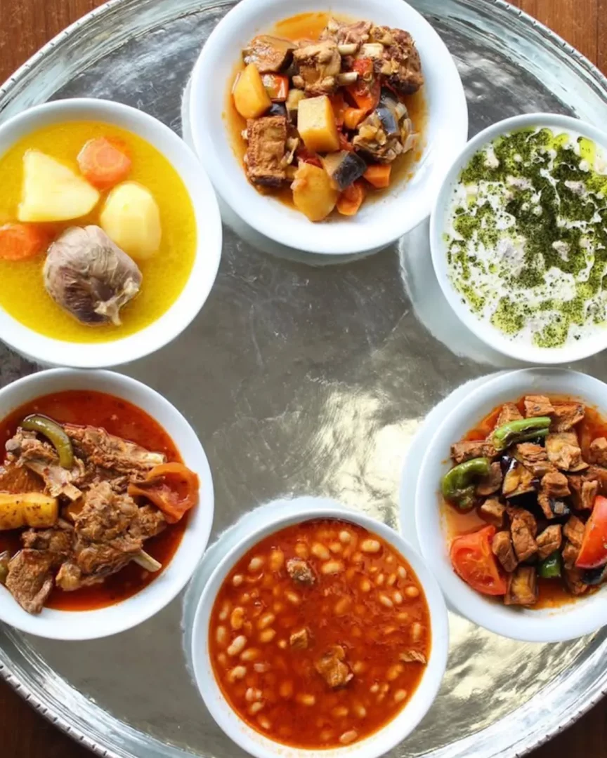 Gümüş bir tepsi üzerinde dairesel olarak dizilmiş altı beyaz kasede; sarımsaklı ve patatesli et çorbası, naneli yoğurtlu meze, zengin soslu kuru fasulye, patlıcanlı ve biberli et güveçleri gibi Gaziantep lokantasına özgü çeşitli geleneksel Türk yemekleri sunuluyor.