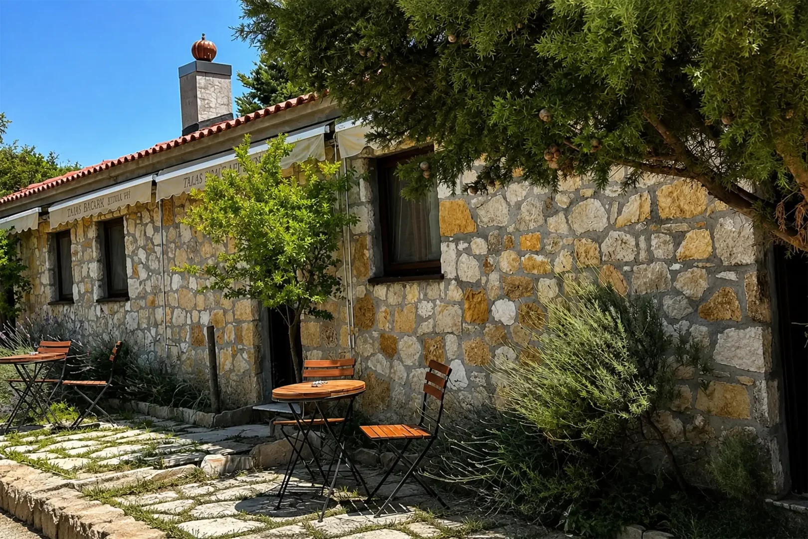 Talay Bağları Konuk Evleri'nden birine ait taş duvarlı, kiremit çatılı bir binanın dış cephesi, önünde ahşap masa ve sandalyelerin bulunduğu, yeşil bitkilerle çevrili, güneşli bir avlu ile bacasındaki balkabağı şeklinde süslemeyi gösteriyor.