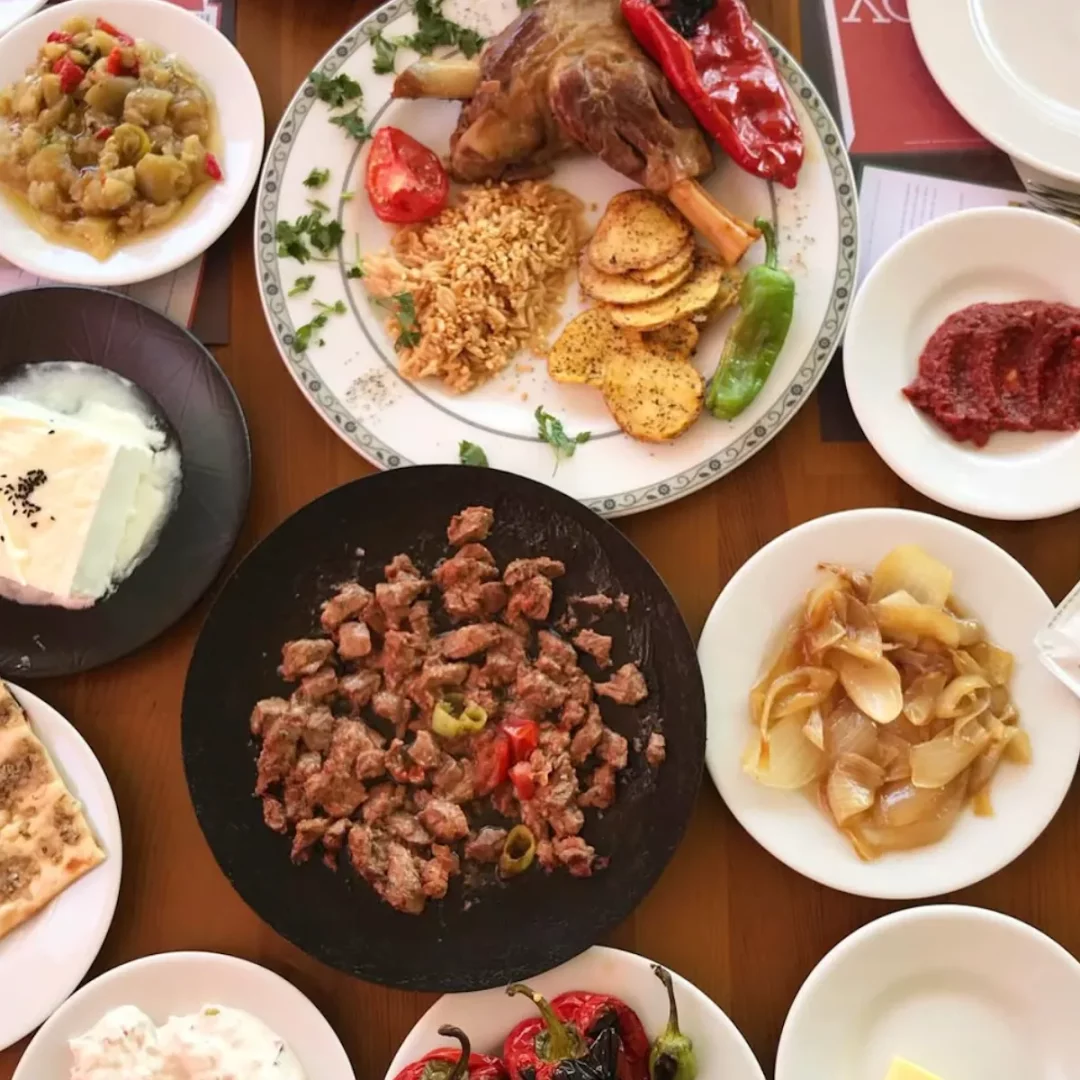 Türk mutfağının zenginliğini yansıtan, üzerinde kemikli et yemeği, pilav, kavrulmuş patates dilimleri ve biberlerin bulunduğu büyük bir tabak, ortaya yerleştirilmiş bol etli tava yemeği, patlıcan salatası, peynir ve yoğurt, acılı ezme ve kavrulmuş soğan gibi çeşitli mezelerin olduğu, Tavacı Refik menüsünden geleneksel bir ziyafet sofrası üstten çekilmiş geniş açılı bir fotoğrafta görülüyor.