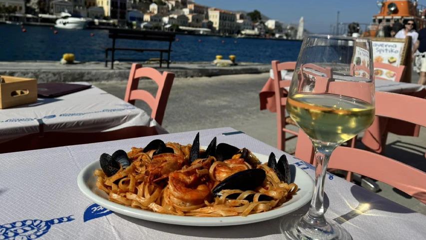 Symi Adası'ndaki Taverna To Spitiko'da, masanın üzerinde iştah açıcı midye ve karideslerle süslü bir deniz mahsulleri makarnası tabağı ve serinletici bir kadeh beyaz şarap, arkada masmavi deniz ve liman manzarası eşliğinde Akdeniz lezzetini sunuyor.