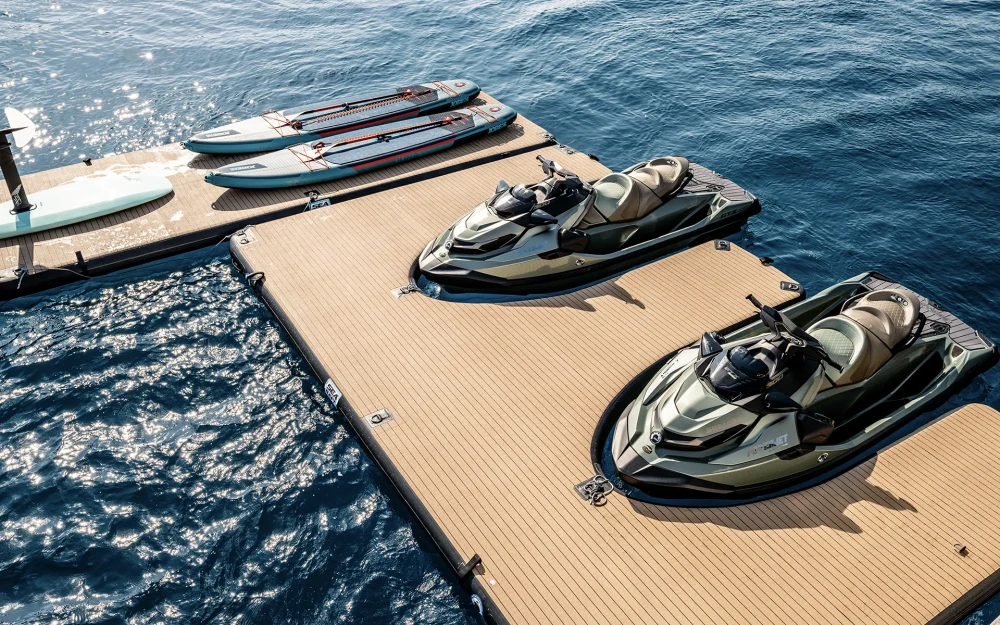 Parlak güneşli bir günde, masmavi denizin üzerinde yüzen kahverengi ahşap desenli geniş şişme platformda, lüks bir tatil deneyimi sunan iki şık jet ski ve birkaç farklı su oyuncağı, kullanıma hazır bekliyor.
