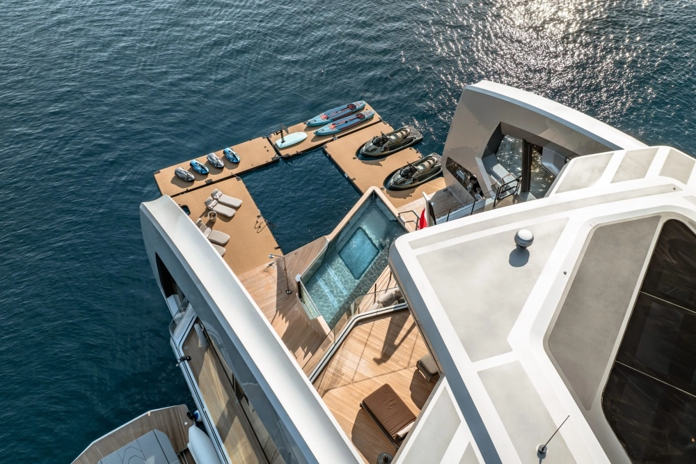Modern bir süper yatın kıç güvertesinden yukarıdan çekilen bu fotoğrafta, şezlonglar, iki jet ski ve çeşitli paddleboard su oyuncaklarının bulunduğu, denize açılan geniş yüzer platform ve yatın güvertesindeki şeffaf yüzme havuzu, lüks bir charter deneyimi sunan detaylarla göz kamaştırıyor.