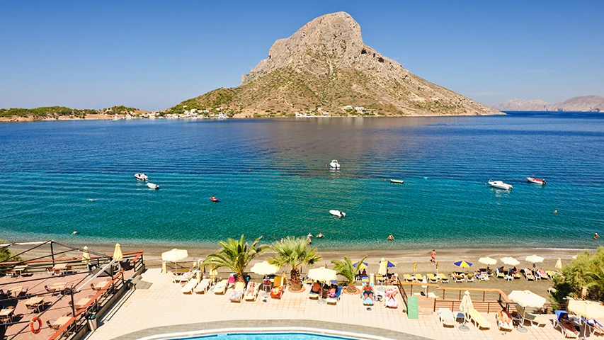 Kalymnos'ta, güneşli bir havada, berrak turkuaz denizin kıyısındaki bir otelin plaj ve havuz alanından Telendos Adası'na doğru uzanan manzarada, denizde yüzen tekneler ve insanlar ile sahilde şezlonglarda dinlenen tatilciler dikkat çekiyor.
