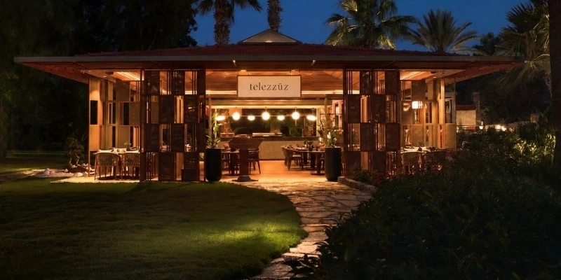 Bodrum'da bulunan, modern ahşap tasarımı ve sıcak aydınlatmasıyla dikkat çeken Telezzüz restoranının gece görüntüsü, yemyeşil çimler ve taş patikayla çevrili dış alanları ve içinde birçok masa ile palmiye ağaçlarını gösteriyor.