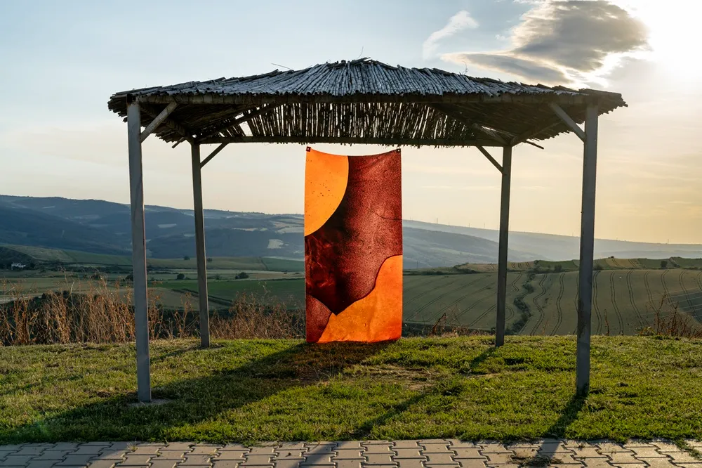 Gün batımının kızıl ışıklarıyla aydınlanan, yemyeşil tepeler ve tarlalarla dolu geniş bir manzaranın önünde, saz çatılı bir gölgeliğin altında asılı, turuncu ve kırmızımsı kahverengi tonlarda soyut organik formlar içeren Temas Bölgesi sergisinden dikkat çekici bir sanat eseri.