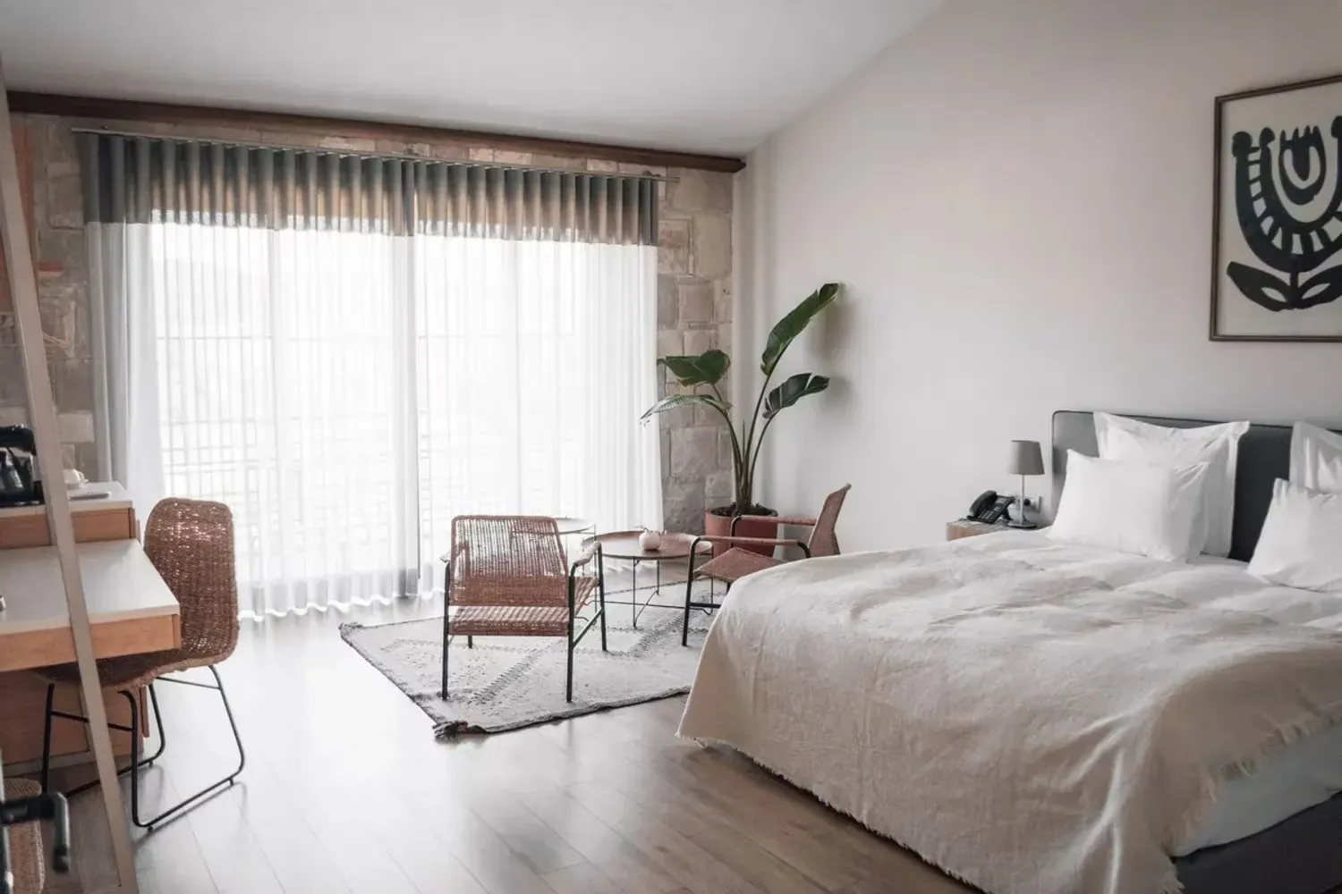 Urla'daki bir otel odasının aydınlık ve modern iç tasarımı; beyaz çarşaflı geniş bir yatak, hasır sandalyeler ve sehpadan oluşan oturma alanı, büyük pencereler, duvarı süsleyen soyut tablo ve doğal bitkilerle huzurlu bir atmosfer sunuyor.