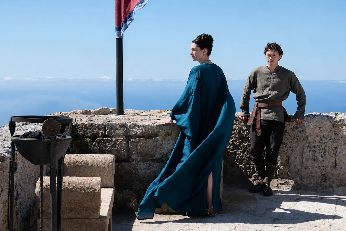 Christopher Nolan'ın The Odyssey filmi kadrosundan Anne Hathaway'in canlandırdığı Penelope, dalgalanan teal rengi elbisesiyle, ve Tom Holland'ın canlandırdığı Telemakhos, sade kıyafetleriyle, Homeros'un epik anlatısındaki bekleyişi ve denize karşı özlemi yansıtan bir sahneyi kalenin taş duvarından engin mavi deniz ve gökyüzüne bakarak sergiliyorlar.