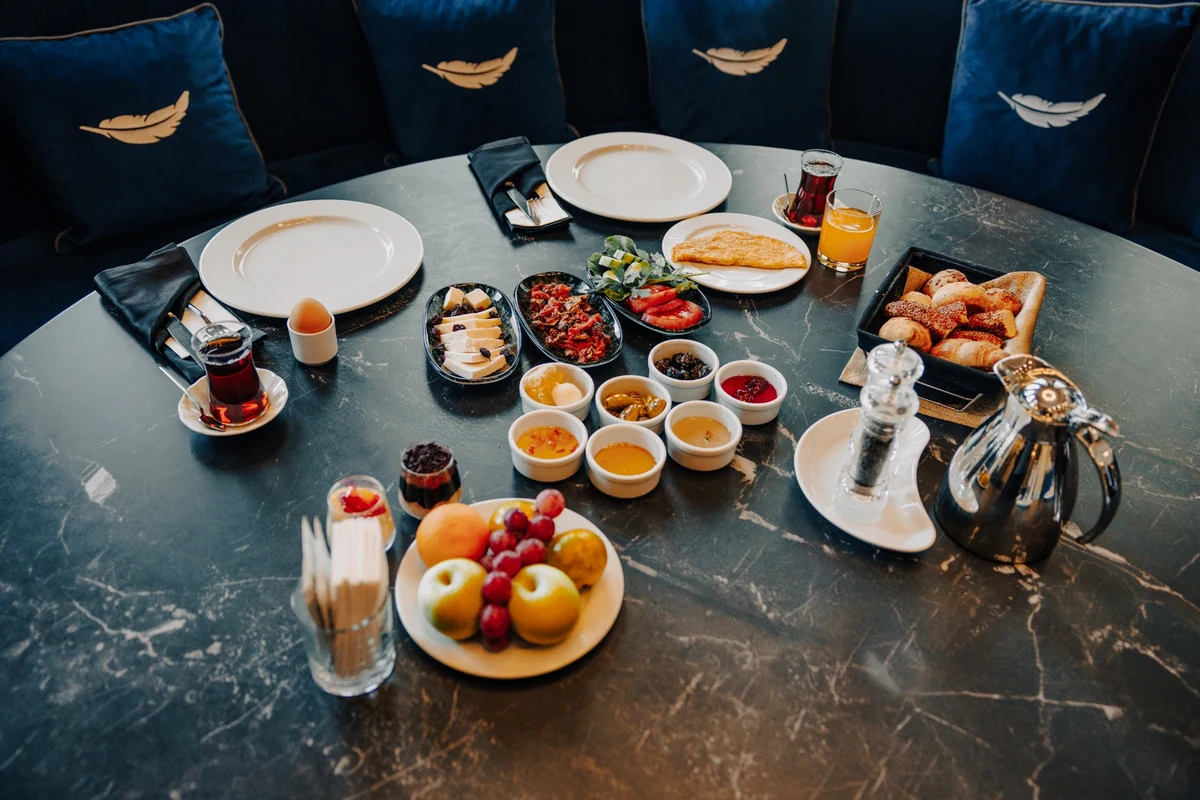 The Wings Hotels Ankara'da mermer bir masanın üzerinde, arkada tüy detaylı lacivert minderlerin bulunduğu, çeşitli peynirler, zeytinler, bal, reçel, taze omlet, sıcacık hamur işleri ve meyvelerle dolu, eksiksiz bir kahvaltı sofrası sergileniyor.