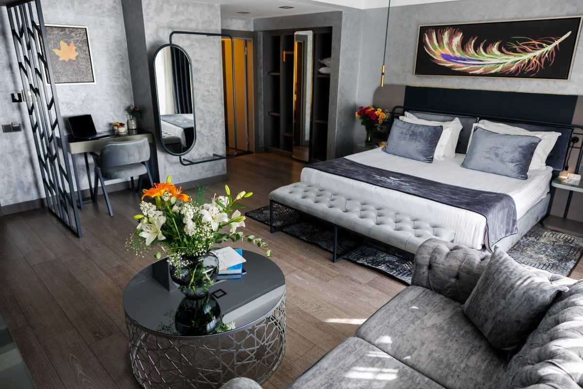The Wings Hotels Ankara adlı oteldeki bu şık ve modern oda, gri tonlarda yumuşak yatak takımlarına sahip büyük bir yatak, ayak ucunda döşemeli bir bank, rahat gri bir kanepe, taze çiçeklerle süslü cam bir sehpa, dizüstü bilgisayarlı çalışma alanı ve duvarda tüylü sanat eseri ile konforlu bir konaklama deneyimi sunuyor.