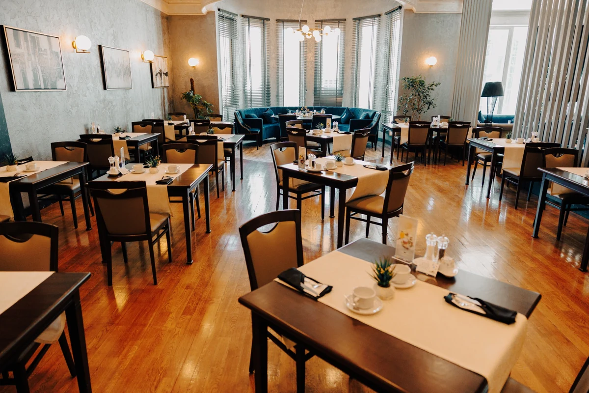The Wings Hotels Ankara restoranının zarif iç mekanı, misafirler için hazırlanmış açık renkli masa örtüleri, fincanlar ve küçük bitkilerle donatılmış çok sayıda yemek masasını ve cilalı ahşap zemini sergiliyor; arka planda jaluzili büyük pencereler ve kavisli mavi koltuklu oturma alanı, aydınlık ve davetkar bir atmosfer yaratıyor.
