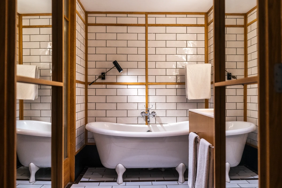 The Wings Hotels Collection'a ait bu zarif banyoda, beyaz metro fayanslarla kaplı duvarlar arasında ahşap çerçevelerle çevrili, ortasında modern siyah aplik ve krom bataryalı ayaklı beyaz bir küvet yer alırken, sağda ahşap bir bölme arkasında benzer bir küvet ve havlular dikkat çekiyor.