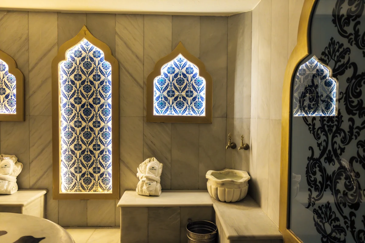 The Wings Hotels Collection'ın hamamında, mermer duvarlar arasında, mavi ve beyaz İznik çinileriyle süslü, altın çerçeveli, aydınlatmalı kemerli paneller, pirinç musluklu geleneksel mermer bir kurna ve The Wings Hotels logosu taşıyan katlanmış bornozlar ile zarif ve otantik bir Türk hamamı atmosferi görülmektedir.