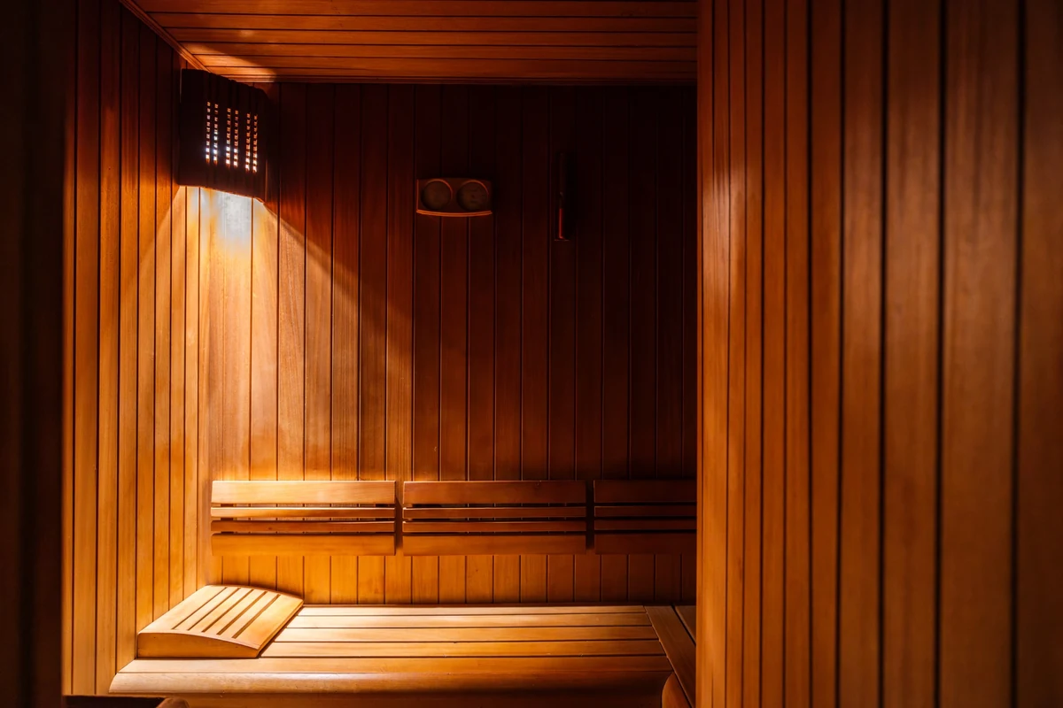 Sıcak ahşap panellerle kaplı, loş ve davetkar ışıkla aydınlatılmış, rahatlatıcı bir sauna odasının iç görünümü, konforlu ahşap bankları ve duvardaki sıcaklık ölçerleriyle huzurlu bir dinlenme ortamı sunuyor.