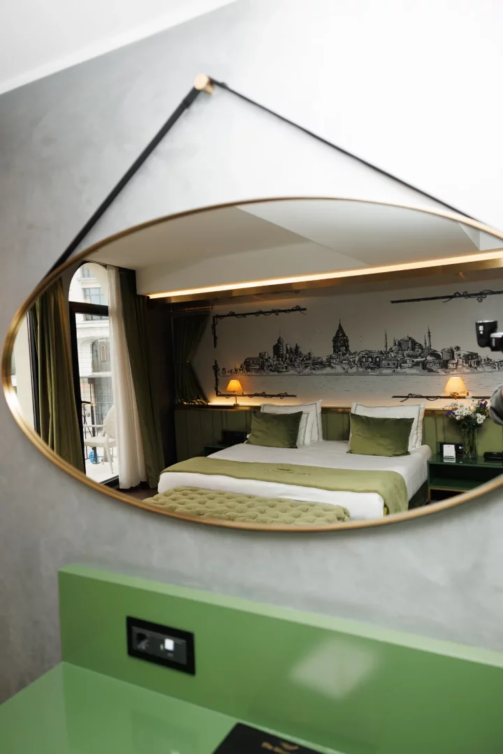 Karaköy'deki The Wings Hotels'in bir odasındaki şık oval ayna, yeşil tonlarında nevresimli konforlu bir yatağı, arka duvarda İstanbul silüeti çizimi ve doğal ışık alan bir balkonu yansıtarak mekanın ambiyansını gözler önüne seriyor.