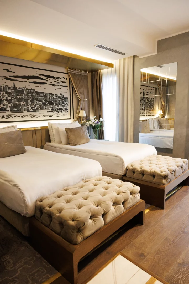 The Wings Hotels Karaköy'deki modern ve davetkar iki yataklı oda, duvarda asılı siyah beyaz şehir manzarası tablosunun altında yer alan iki tek kişilik yatak, yatakların ayakucundaki kadife kapitone puflar ve genişlik hissi veren şık bir aynalı duvarla konforlu bir konaklama alanı sunmaktadır.