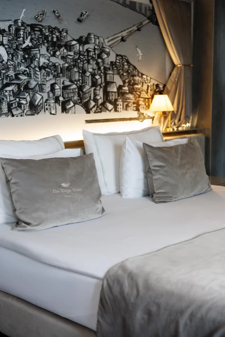 Ortada bembeyaz çarşaflarla özenle hazırlanmış bir yatak, başucunda The Wings Hotel logolu gri kadife yastıklar ve arka planda Galata Köprüsü ile Haliç'i andıran siyah beyaz şehir manzarası çizimi, otelin konforlu ve eşsiz Karaköy odasını sergiliyor.