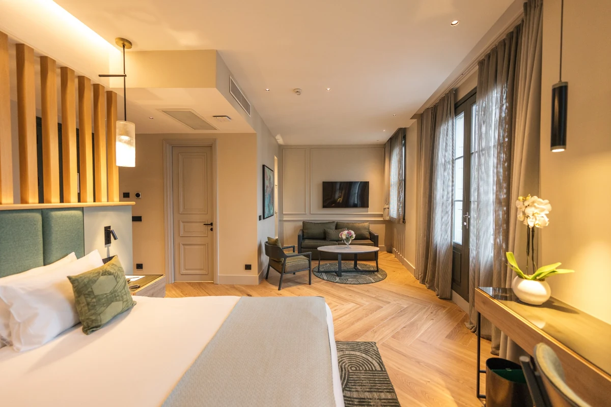 The Wings Hotels Pera geniş suite odanın yatak ve oturma alanlarını gösteren bu görüntüde, yeşil yatak başlığı ve örtülü konforlu bir yatak, ahşap zemin, duvara monte televizyonlu koyu yeşil bir koltuk takımı ve büyük pencerelerle aydınlık bir yaşam alanı dikkat çekiyor.