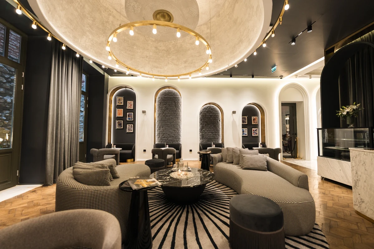 The Wings Hotels Pera lobisinde, siyah-beyaz kazayağı desenli kıvrımlı koltukların ve ışınsal çizgili halının hakim olduğu, kemerli nişler, ahşap zemin ve altın rengi modern aydınlatmalarla donatılmış, konforlu ve şık bir oturma alanı sergileniyor.