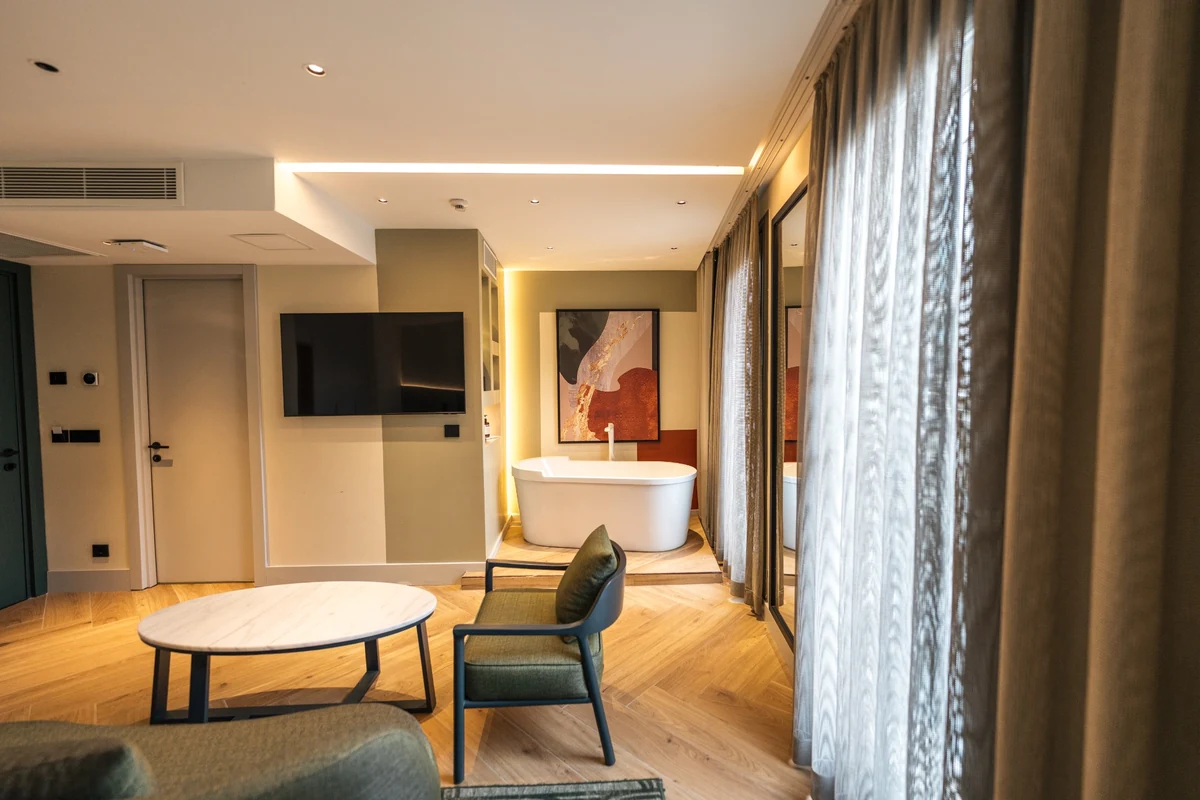 The Wings Hotels Pera süitindeki modern yaşam alanıyla bütünleşen, duvara monte televizyon, şık mermer sehpa ve koyu yeşil koltuk takımı içeren bir köşe ile ayaklı beyaz küvetin bulunduğu, parke zeminli ve gün ışığı alan pencereli lüks bir banyo görünümü.