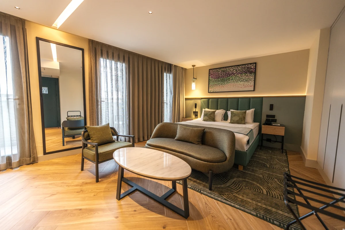 The Wings Hotels Pera süit odası, yeşil yatak başlığı ve uyumlu yatak takımlarıyla donatılmış geniş çift kişilik yatağı, rahat yeşil koltuk ve kanepesi, oval mermer sehpası ve doğal ışığı içeri alan pencereleriyle modern ve konforlu bir atmosfer sunuyor.