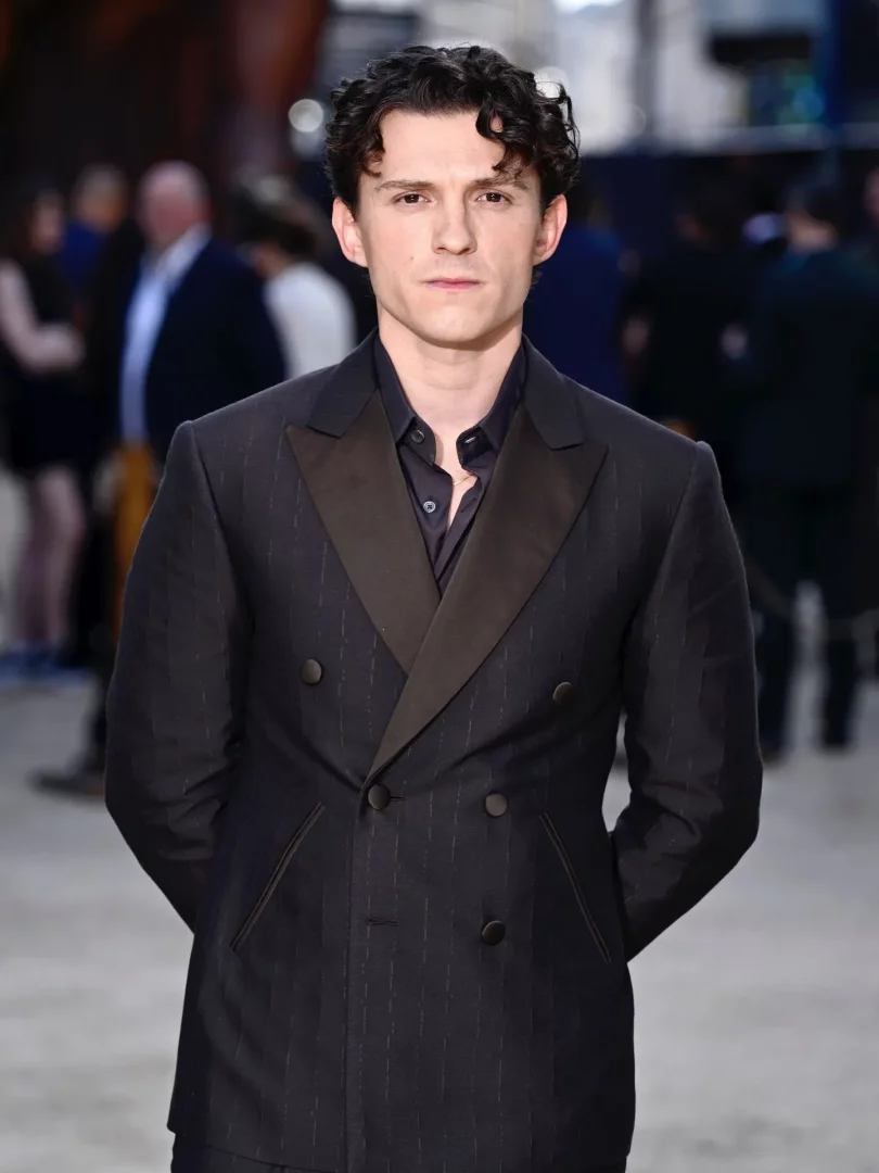 Tom Holland, The Odyssey dünya prömiyerinde Gieves & Hawkes marka lacivert çizgili kruvaze takım elbisesi, parlak koyu renk şal yaka detayı ve koyu renk gömleğiyle, elleri arkasında duran, kıvırcık saçlı genç bir adam olarak doğrudan kameraya bakıyor.
