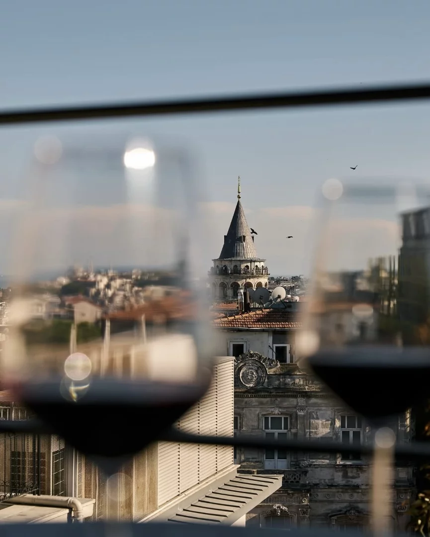 Galata Kulesi'nin siluetinin iki kadeh kırmızı şarap arasından görüldüğü, Asmalımescit'ten parlak güneşli bir havada İstanbul'un çatılarını ve şehir manzarasını yansıtan büyüleyici bir görüntü.