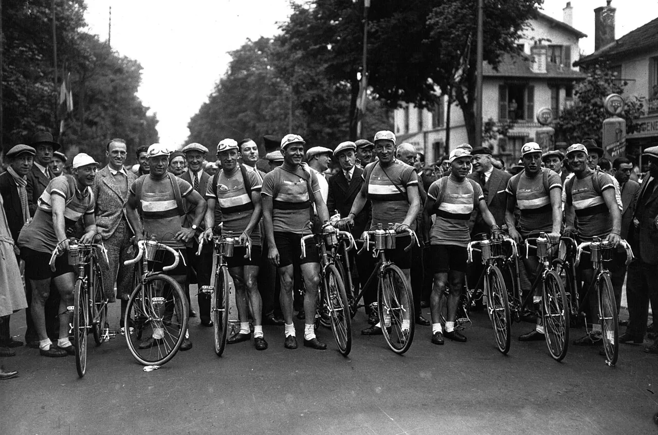 1932 Tour de France bisiklet yarışından siyah beyaz bu tarihi fotoğrafta, çizgili formalar ve kask benzeri şapkalar giymiş bir grup erkek bisikletçi, yarış öncesi bisikletleriyle ağaçlarla çevrili bir bulvar üzerinde sıralanmış, arkalarında kalabalık izleyiciler görülüyor.