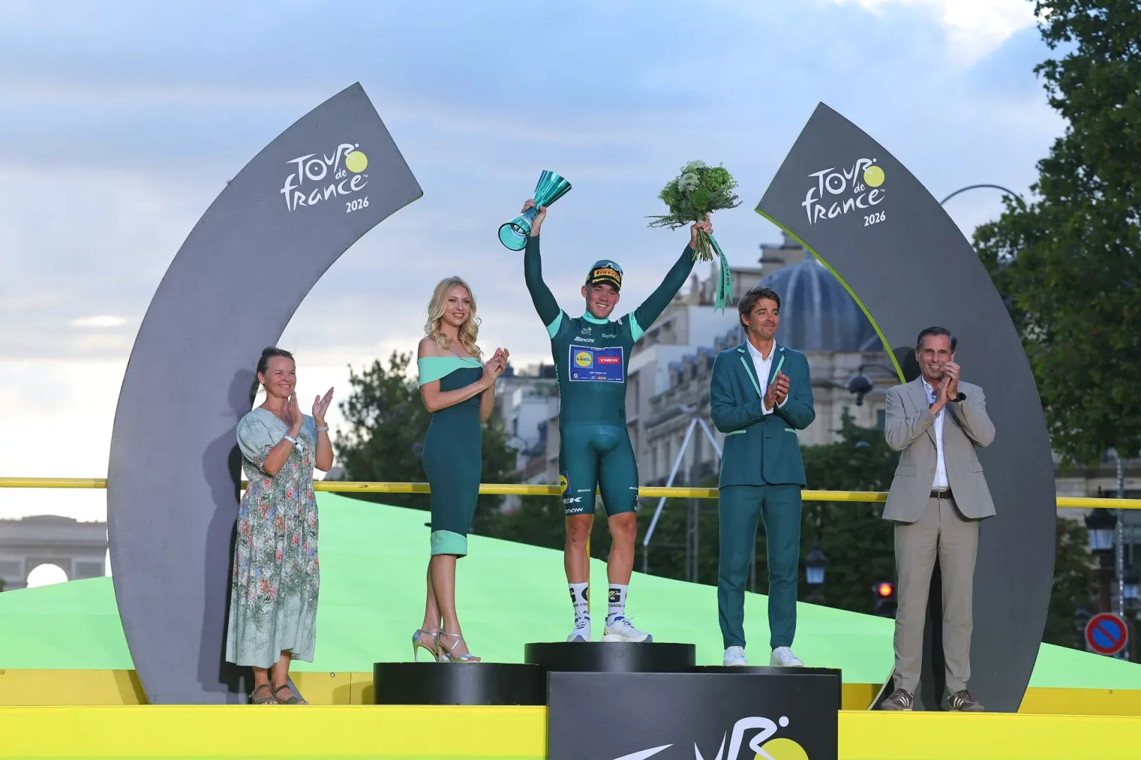 Tour de France 2026 ödül podyumunda, yeşil bisikletçi forması içindeki bir sporcu, kupasını ve çiçek buketini coşkuyla havaya kaldırarak zaferini kutlarken, alkışlayan dört kişi de bu önemli anı onunla birlikte paylaşıyor, arka planda şehrin mimarisi hafifçe seçiliyor.