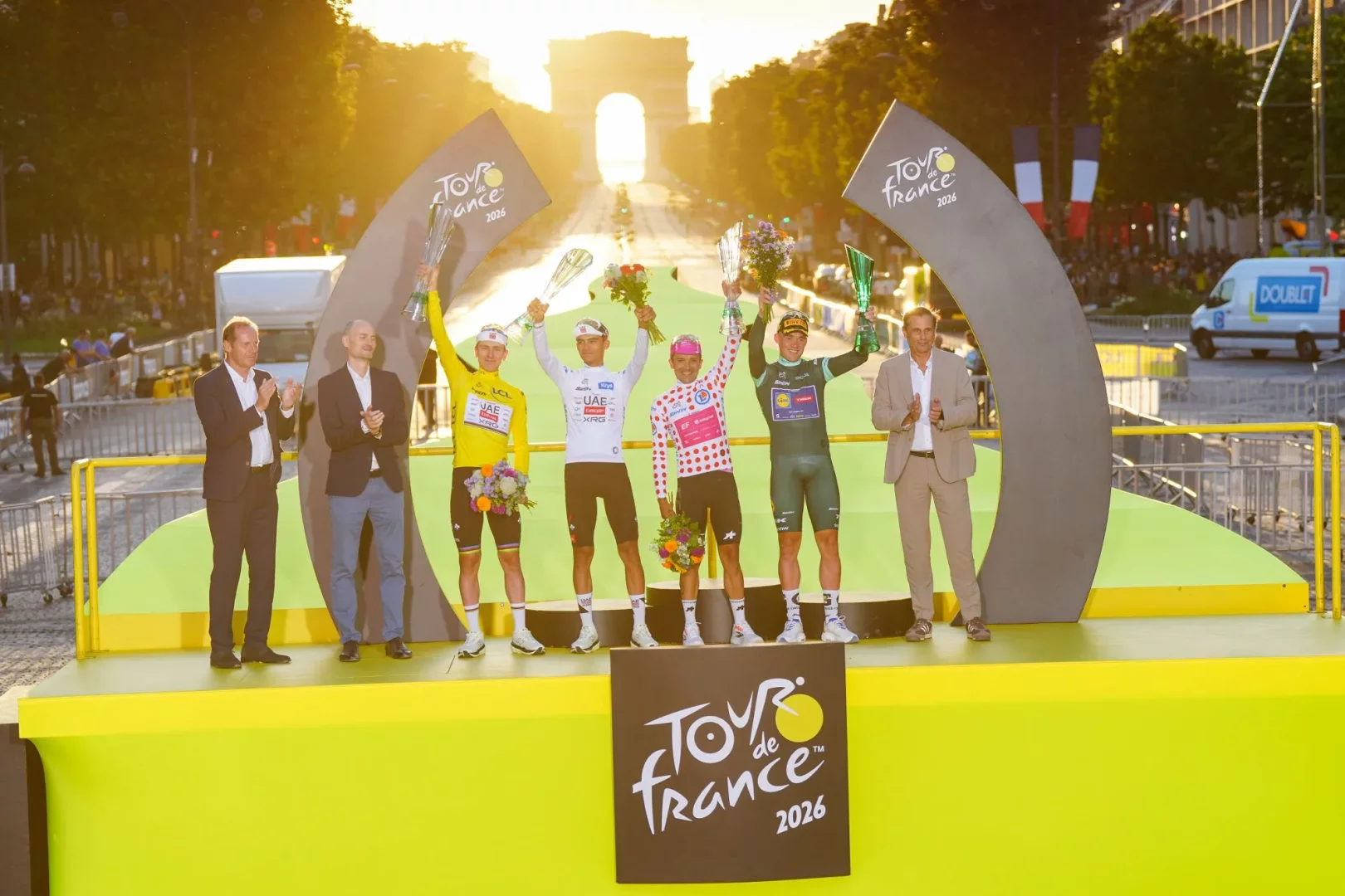 Güneşin Arc de Triomphe'un arkasından güçlü bir ışıkla aydınlattığı görkemli bir Tour de France 2026 ödül töreni sahnesinde, sarı, beyaz, puantiyeli ve yeşil mayolarıyla podyumdaki dört bisikletçi coşkuyla kupalarını ve çiçek buketlerini havaya kaldırırken, iki takım elbiseli adam da onları alkışlıyor.