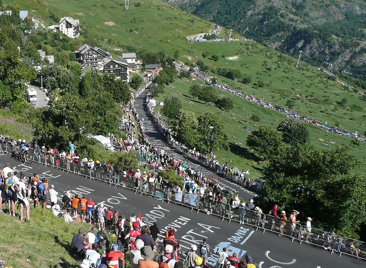 Tour de France Alpe d'Huez etabında, virajlı dağ yolunun kenarında toplanmış binlerce seyirci, yokuş yukarı ilerleyen bisikletçileri çitlerin arkasından ve yeşil yamaçlardan coşkuyla destekliyor; asfalt üzerindeki yazılar ve dağlık çevredeki binalar o anın atmosferini güçlendiriyor.