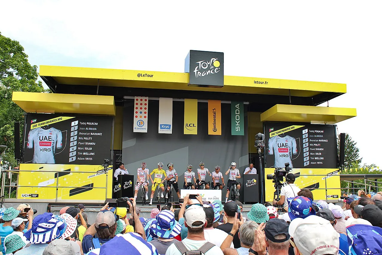 Tour de France etkinliğinde, parlak sarı bir podyumda dizilmiş bisikletçiler, dev ekranlarda UAE Team Emirates XRG takımının forması ve Tadej Pogačar liderliğindeki derece listeleri gösterilirken, sponsor logolarının altında coşkulu kalabalıklar bu anları fotoğraflıyor, bu da yarışmanın puanlama sistemi ile ilgili bilgileri yansıtıyor.
