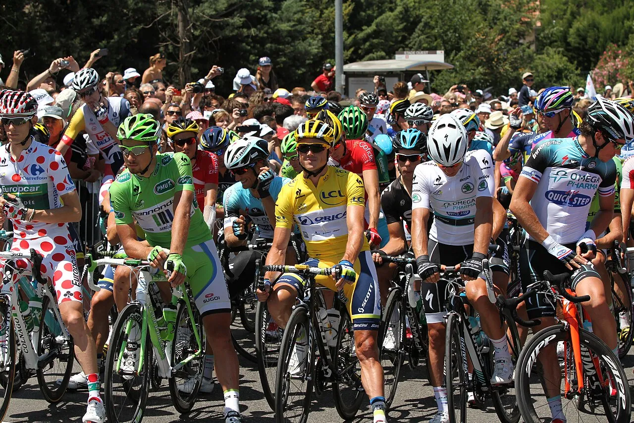 Bir Tour de France yarışının başlangıcında, sarı, yeşil, beyaz puantiyeli ve beyaz gibi farklı renklerde mayolar giyen bisikletçiler, arkalarında fotoğraf çeken kalabalık seyircilerle birlikte yarışa hazır bir şekilde duruyorlar.