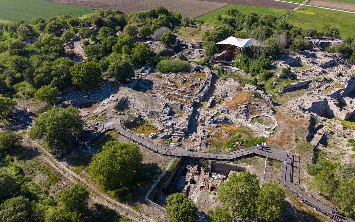Çanakkale'deki Troya Antik Kenti'nin yemyeşil ağaçlar ve uzaktaki tarım arazileriyle çevrili, kısmen kazılmış taş yapı kalıntıları, antik tiyatro bölümü ve üzerlerinde gezinen ziyaretçilerin bulunduğu modern ahşap yürüyüş yolları ile beyaz koruyucu bir çadırın görüldüğü kuşbakışı genel görünümü.
